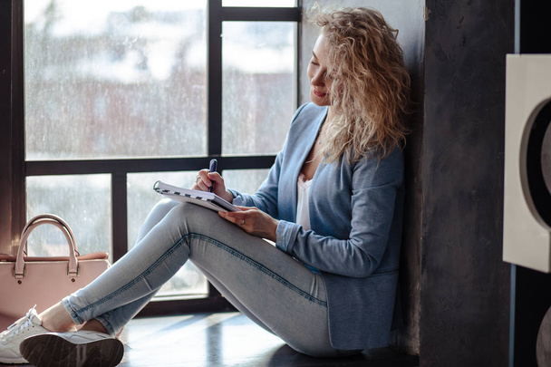Pretty blonde young business woman sitting near window with planner notebook. Lifestyle casual portrait - Foto, imagen