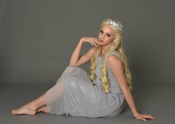 full length portrait of blonde woman wearing crown and pale blue dress. seated pose against a grey studio background. - Фото, изображение