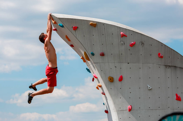 Photo of strong sports guy in red shorts hanging on wall for climbing against blue sky with clouds - Foto, Bild