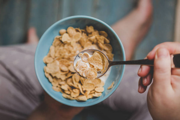 Woman hold sky blue colored bowl of tasty cereal corn flakes with jug of milk pouring into bowl on a wooden surface from above - Fotografie, Obrázek