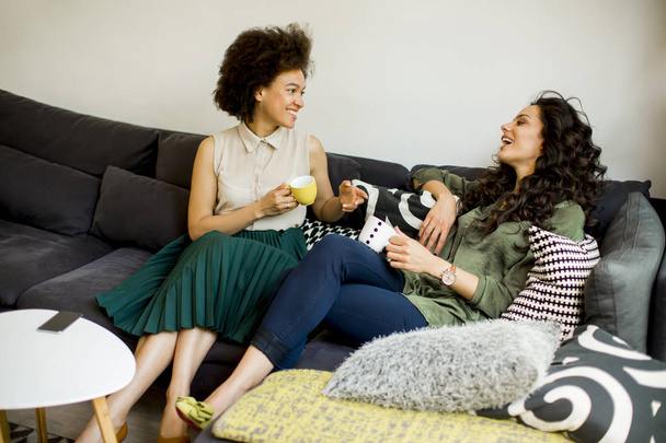 Kaksi monirotuista nuorta naista juttelemassa ja juomassa kahvia olohuoneessa
 - Valokuva, kuva