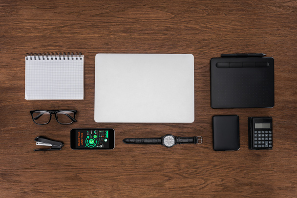 top view of workplace with arranged empty textbook, laptop, wristwatch and smartphone with marketing analysis on screen - Zdjęcie, obraz