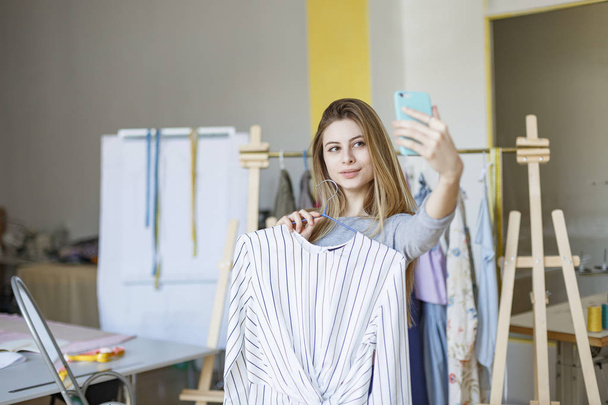 Chica bonita sosteniendo nuevo vestido en la percha en la mano tomando fotos en la cámara frontal del teléfono celular en el taller de costura moderno
 - Foto, imagen