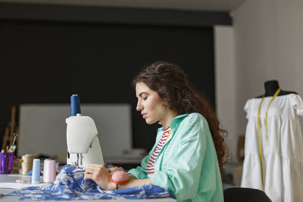Joven costurera en camisa colorida cuidadosamente trabajando con la máquina de coser en taller de costura moderno
 - Foto, Imagen