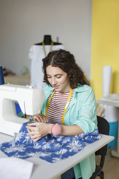 Красивая улыбающаяся швея в красочной рубашке и полосатой футболке мечтательно работающая на швейной машинке в современной швейной мастерской
 - Фото, изображение