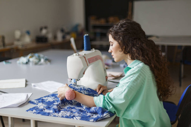 Jovem costureira com cabelos encaracolados escuros em camisa colorida cuidadosamente usando máquina de costura na oficina de costura moderna
 - Foto, Imagem