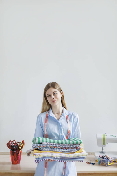 junges schönes Mädchen in blauem Hemd schaut glücklich in die Kamera und hält Muster Stoffe in den Händen in der Nähwerkstatt - Foto, Bild