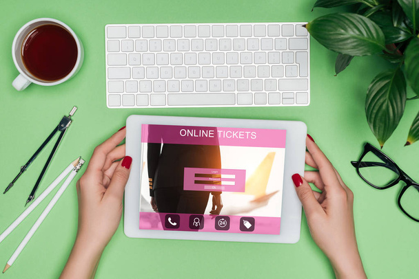bijgesneden afbeelding van vrouwelijke architect digitale tablet met online tickets op scherm aan tafel met de scheidingslijn, koffie en ingemaakte planten houden  - Foto, afbeelding