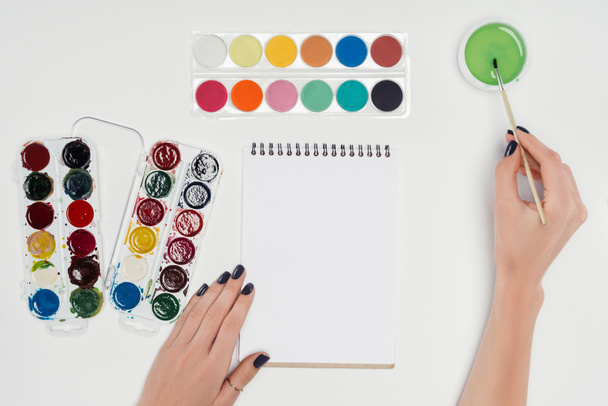 обрезанное изображение художницы, рисующей кистью в пустом учебнике за белым столом с красочными красками
  - Фото, изображение