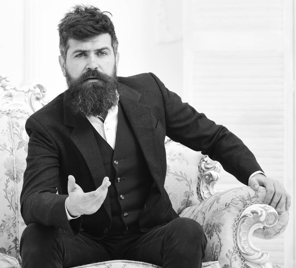 Man with beard and mustache wearing classic suit, sits on old fashioned armchair or sofa. Macho attractive and elegant on serious face and explaining expression. Speaker, wise lecturer concept - Valokuva, kuva