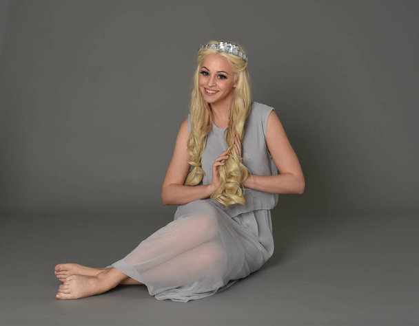Ganzkörperporträt einer blonden Frau mit Krone und blassblauem Kleid. sitzende Pose vor grauem Studiohintergrund. - Foto, Bild