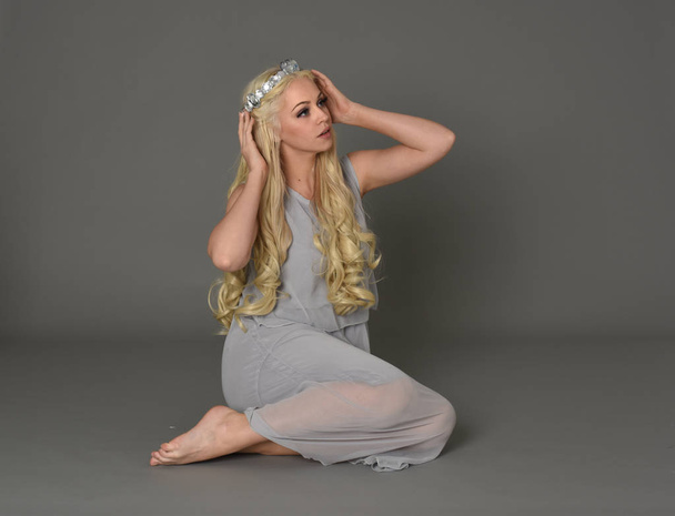 Ganzkörperporträt einer blonden Frau mit Krone und blassblauem Kleid. sitzende Pose vor grauem Studiohintergrund. - Foto, Bild