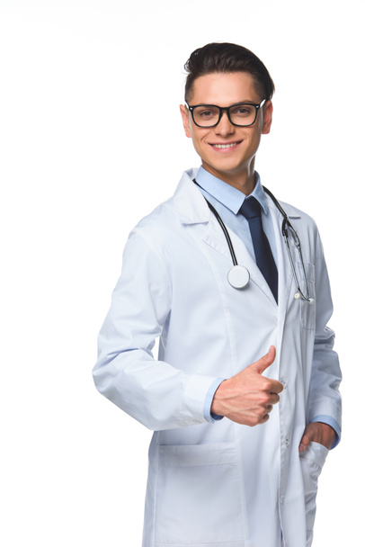 usmívající se mladý lékař ukazuje palcem a při pohledu na fotoaparát izolované na bílém - Fotografie, Obrázek