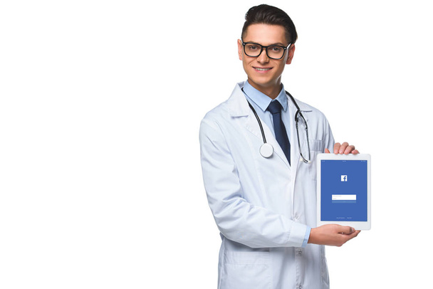 handsome young doctor holding tablet with facebook app on screen isolated on white - Φωτογραφία, εικόνα