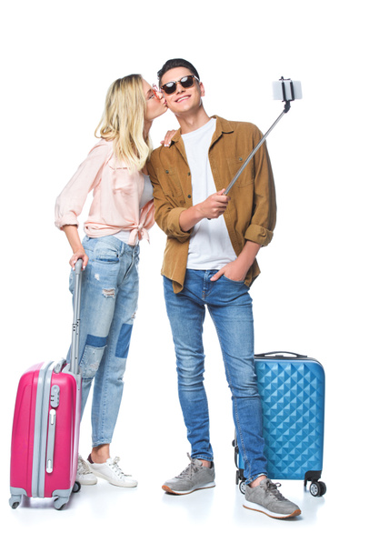 νεαρό ζευγάρι με βαλίτσες λήψη selfie από μονόποδο ενώ γυναίκα φιλώντας το φίλο που απομονώνονται σε λευκό - Φωτογραφία, εικόνα