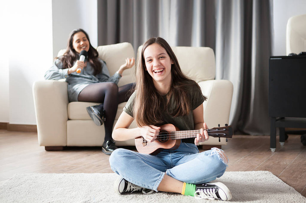 Two sisters, the younger one is playing a small guitar in front at the other is singing in the back - Photo, Image
