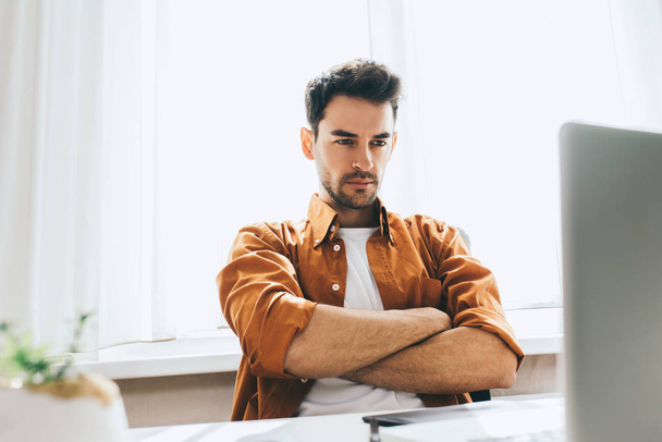 Serious and handsome male freelancer sitting in front of the computer with serious and thoughtful expression crossed his arms. Businessman think about resolves some problems in the office. - 写真・画像