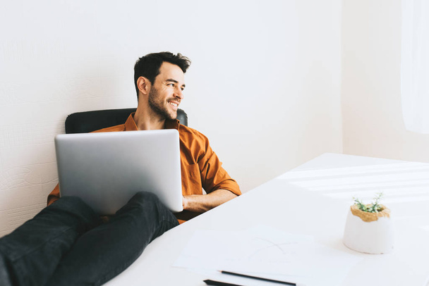 Attractive shot of happy freelancer with cheerful smile achieving life goals. Success and achievement concept. Happy Caucasian businessman sitting next to computer and relax with legs on table. - Fotó, kép