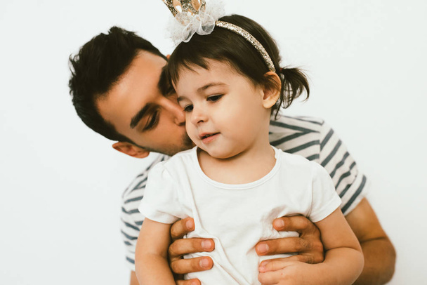 Horizontal close up shot of cute toddler girl wearing crown, on her birthday embrace with dad against white background. Portrait of joyful father kiss and hug his daughter. Happy relationship family. - Photo, Image