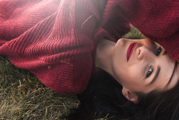 Side view portrait of a beautiful young sexy brunette woman smiling, wearing knitted sweater and looking at the camera, lying on the grass enjoying the weahter. Relaxation, travel and people concept. - Photo, Image