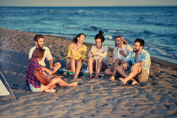 Grupo de amigos sorridentes juntos relaxando com tocar guitarra e cantar uma música na praia do mar
 - Foto, Imagem