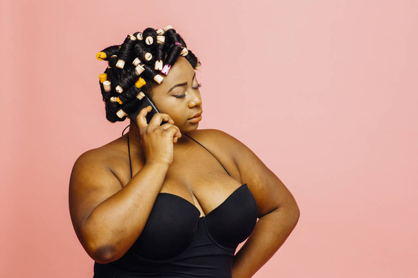 Woman with hair curlers and neglige talking on the phone with eyes closed, isolated on pink background - Photo, image