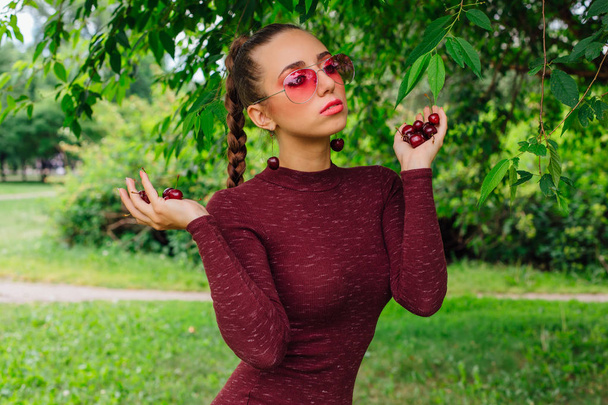 Beautiful young woman with long braid, pink glasses and cherry earrings holding cherry in hands - Fotó, kép