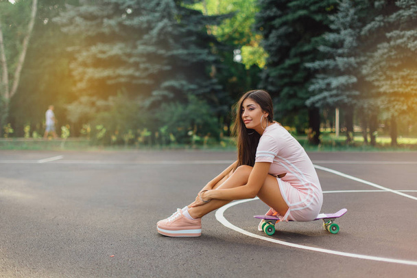 Portrait of a smiling charming brunette female sitting on her skateboard on a basketball court. Happy woman with trendy look taking break during sunset. - Zdjęcie, obraz