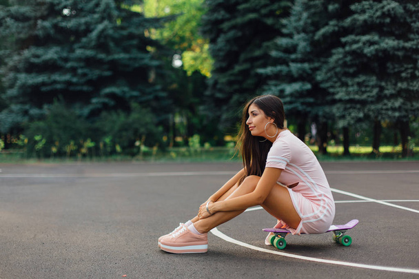 Portrait of a smiling charming brunette female sitting on her skateboard on a basketball court. Happy woman with trendy look taking break during sunset. - Foto, Bild