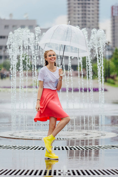 Όμορφη κοπέλα με δύο πλεξούδες σε κίτρινες μπότες και με διάφανη ομπρέλα στέκεται κοντά σε σιντριβάνι. Βροχερή μέρα στη πόλη. - Φωτογραφία, εικόνα