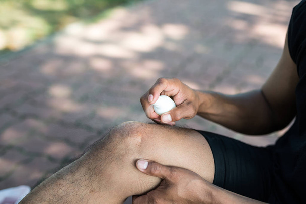 Ein junger Läufer aus Asien mit einer Knieverletzung bekommt Kühlspray zur Linderung der Schmerzen. Konzept der Verletzungen durch das Training. - Foto, Bild