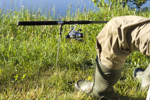 Angler's leg in wellingtons against the meadow and water. Ground rod mounted on a rack - Photo, Image