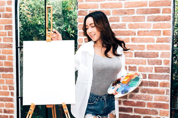 Young woman artist posing with empty canvas and paintbrush at home - Photo, Image