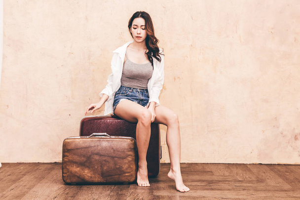 Beautiful traveler woman sit on vintage brown traveling luggage suitcase with wall background - 写真・画像