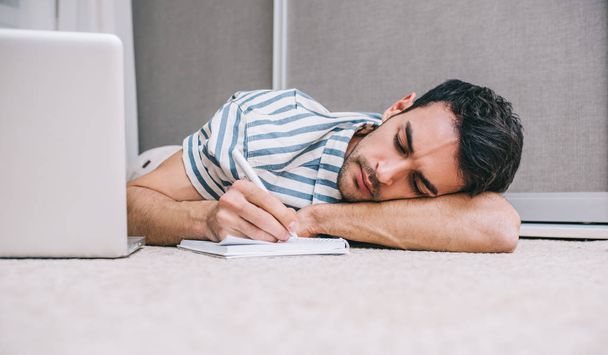 Young Caucasian freelancer business man sleeping near to laptop with eyes closed while lying on the floor home and notice paper. People, lifestyle, technology, business and communication concept. - Foto, immagini