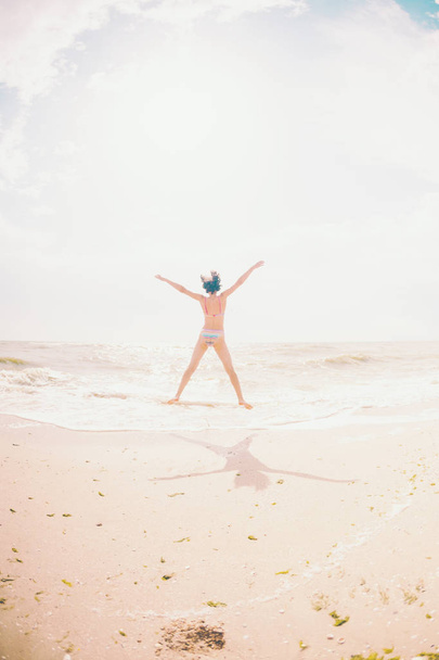 Una ragazza sta saltando sulla spiaggia e mettendo le mani larghe. Una donna snella riposa sulla costa del mare. Bruna sulla riva dell'oceano. Fitness in natura
. - Foto, immagini