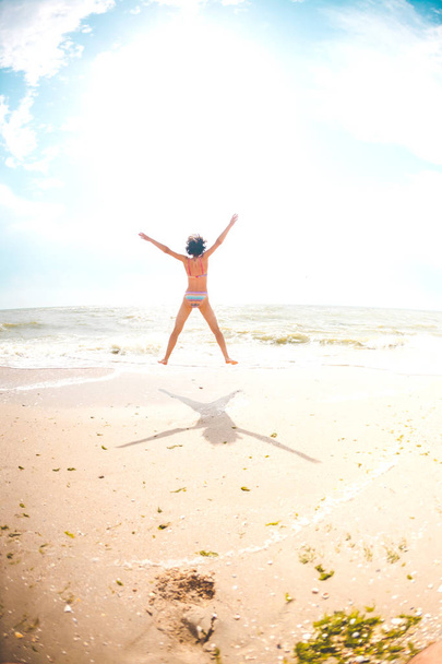 Una ragazza sta saltando sulla spiaggia e mettendo le mani larghe. Una donna snella riposa sulla costa del mare. Bruna sulla riva dell'oceano. Fitness in natura
. - Foto, immagini