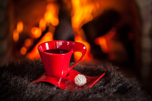 Piros csésze tea vagy kávé és csokoládé, az asztalon a kandalló mellett. Téli és karácsonyi ünnep fogalma. - Fotó, kép