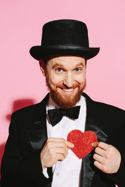 Homem sorrindo em um smoking e chapéu superior segurando um coração vermelho
 - Foto, Imagem