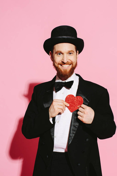 Homem sorrindo em um smoking e chapéu superior segurando um coração vermelho
 - Foto, Imagem