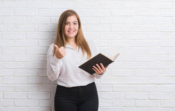 Young adult woman standing over white brick wall reading a book happy with big smile doing ok sign, thumb up with fingers, excellent sign - Zdjęcie, obraz