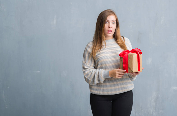 Young adult woman over grey grunge wall holding a present scared in shock with a surprise face, afraid and excited with fear expression - Foto, Imagem