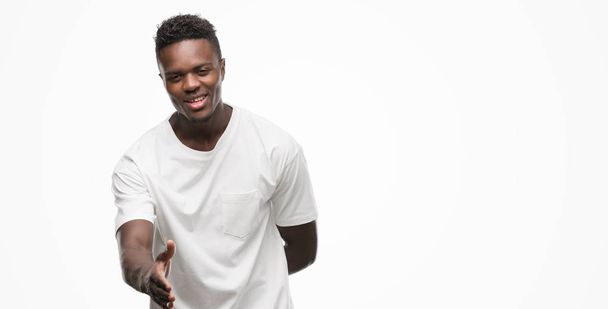 Joven hombre afroamericano vistiendo camiseta blanca sonriendo amigable ofreciendo apretón de manos como saludo y bienvenida. Negocios exitosos
. - Foto, Imagen