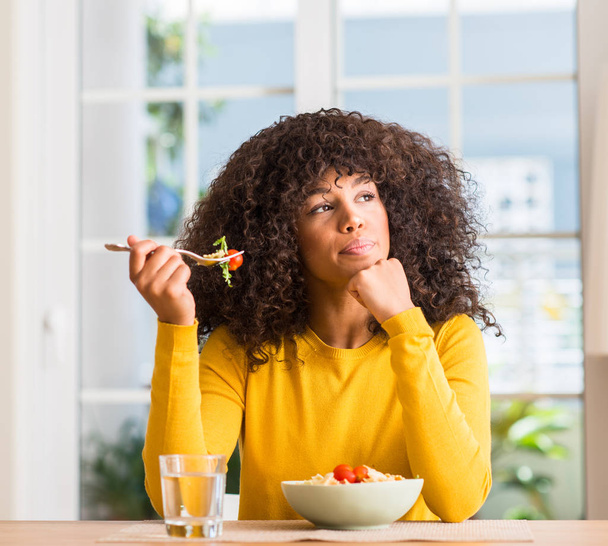 Mulher afro-americana comendo salada de macarrão em casa rosto sério pensando em questão, ideia muito confusa
 - Foto, Imagem