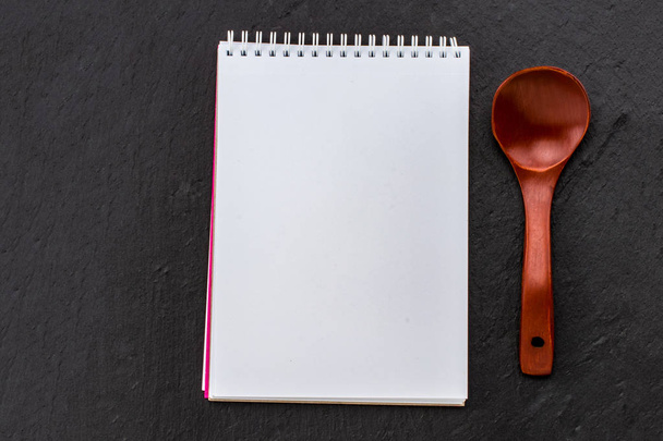 Σημειωματάριο για τη συνταγή σας με ξύλινο κουτάλι πάνω από το μαύρο φόντο πέτρα. Το Top view με αντίγραφο χώρου. - Φωτογραφία, εικόνα