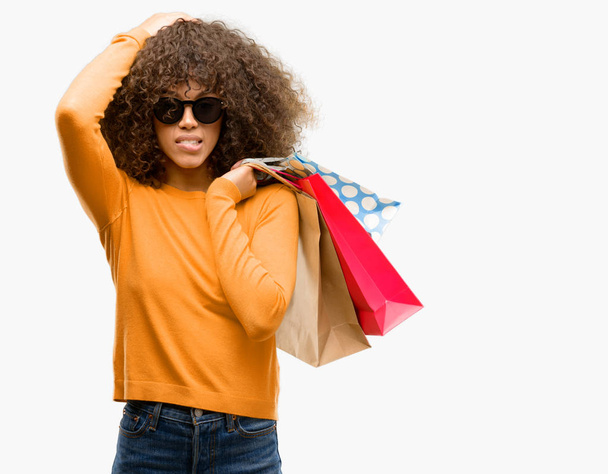 Mujer afroamericana sosteniendo bolsas de compras estresadas con la mano en la cabeza, conmocionadas con vergüenza y cara sorpresa, enojadas y frustradas. Miedo y malestar por error
. - Foto, imagen