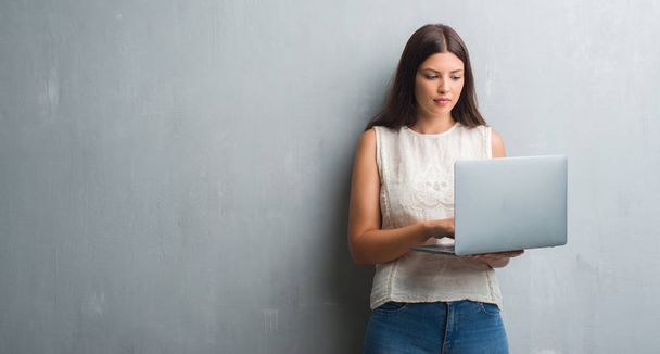 Young brunette woman over grunge grey wall using computer laptop with a confident expression on smart face thinking serious - Photo, Image