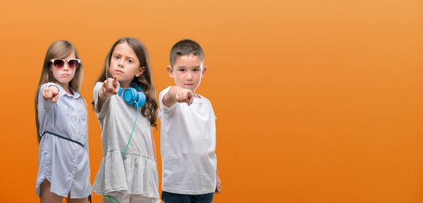 カメラと指で指しているオレンジ色の背景に子供たちの男の子と女の子のグループ, 手に署名、正面からポジティブで自信のジェスチャー - 写真・画像