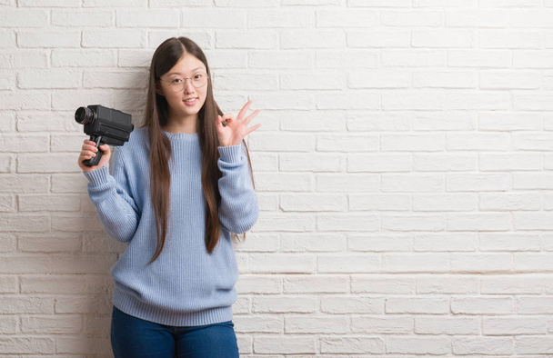 Young Chinese woman filming holding video camera doing ok sign with fingers, excellent symbol - Фото, изображение