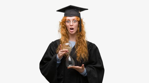 junge rothaarige Studentin in Uniform mit Sanduhr in der Hand erschrocken mit einem Überraschungsgesicht, ängstlich und aufgeregt mit Angstausdruck - Foto, Bild